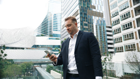 Caucasian business man hold mobile phone and looking at skyscraper view. Skilled male leader standing rooftop in modern building. Caucasian executive manager look at phone while rest his eyes. Urbane.の写真素材