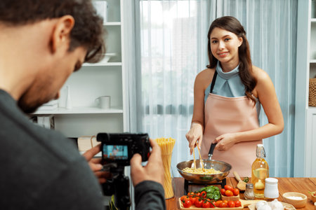 Cameraman recording to woman in chef influencer host cooking spaghetti with meat topped tomato sauce surrounded ingredients recipe, presenting special dish healthy food at modern studio. Postulate.の写真素材
