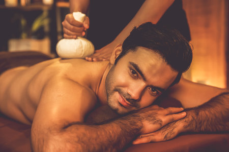 Hot herbal ball spa massage body treatment, masseur gently compresses herb bag on man body. Tranquil and serenity of aromatherapy recreation in warm lighting of candles at spa salon. Quiescentの写真素材