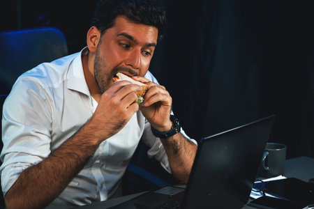 Businessman enjoy eating with delicious sandwich dish on desk while staring laptop at night time. Hungry creative checking email while having a bread meal at workplace with neon blue light. Sellable.の写真素材
