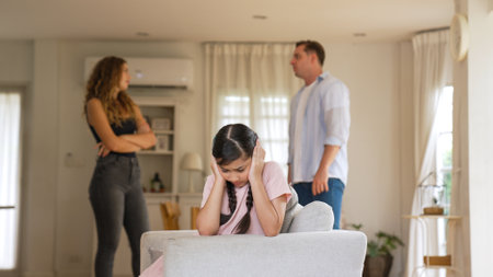 Annoyed and unhappy young girl sitting on sofa trapped in middle of tension by her parent argument in living room. Unhealthy domestic lifestyle and traumatic childhood develop to depression Synchronosの写真素材