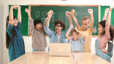 Teacher and multicultural student looking at laptop while celebrate success project and put hands in the air. Group of children smiling to camera while standing at blackboard in classroom. Pedagogy.の写真素材