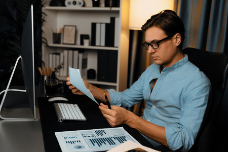 Thinking smart businessman holding paperwork report paragraph dynamic database with pen for analyzing to present customer target for company meeting at modern home office at night time. Pecuniary.の写真素材