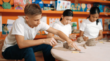 Highschool teenager using carving tool working at clay at pottery workshop. Group of happy diverse children working or modeling cup of clay with dough and equipment scatter around. Edification.の写真素材