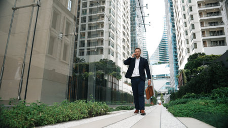 Low angle camera view of caucasian businessman calling to manager and hold suitcase while walking at skyscraper view. Skilled project manager working on telephone and sharing marketing ideas. Urbane.の写真素材