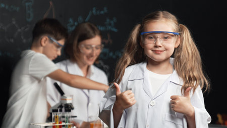 Smart caucasian girl showing thumb while teacher and student doing experiment behind. Child looking at camera while boy looking under microscope at black board written chemical theory. Erudition.の写真素材