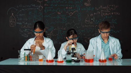 Cute girl looking under microscope while student doing experiment at blackboard with theory written. Young scientist inspect colored solution at table with experimental equipment placed. Edification.の写真素材