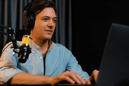 Host channel smart broadcaster talking on live social media application searching on laptop, broadcasting streamer wearing headphone using mic at modern home office studio in warm lighting. Pecuniary.の写真素材