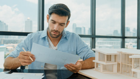 Portrait of smart skilled engineer checking house model while holding blueprint. Professional...