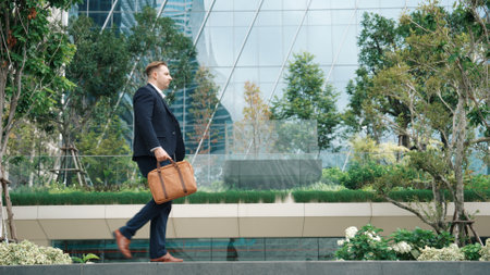 Side view of caucasian businessman hold suitcase and walking at modern building in green eco city. Professional male leader going to workplace at morning while wearing formal suit. Lifestyle. Urbane.の写真素材