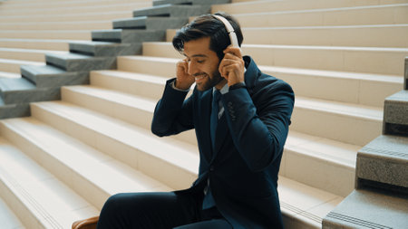 Project manager listening and enjoy music from headphone while sitting at stairs. Professional businessman wearing suit outfit while dancing and moving to music. Happy man listen funny song. Exultant.の写真素材