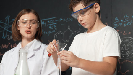 Close up of caucasian boy mixing chemical liquid while teacher giving advice. Instructor wearing lab...