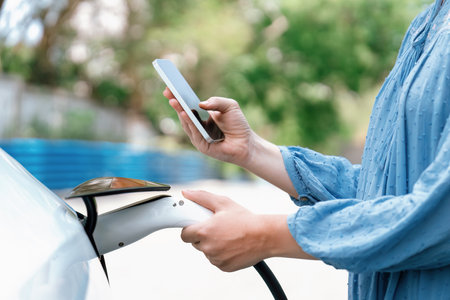 Hand insert EV charger plug into electric vehicle to recharge EV car, battery status display on smartphone EV application. Future alternative clean and sustainable energy for transportation. Perpetualの写真素材