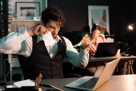 Sitting man with serious face tearing failure paperwork of business project at night lighting time at modern office with blurry secretary checking paper report with folder background. Postulate.の写真素材