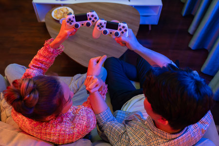 Top view of hands young couple gamer with crashing joysticks, holding hands be winner together, sitting sofa playing video game at back side at neon light color living room at modern home. Infobahn.の写真素材