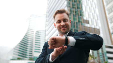 Low angle camera of businessman looking at watch to remind about time. Smart caucasian businessman checking time for being on time in important appointment or meeting with modern skyscraper. Urbane.の写真素材