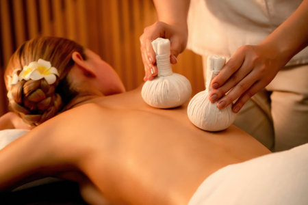 Hot herbal ball spa massage body treatment, masseur gently compresses herb bag on woman body. Tranquil and serenity of aromatherapy recreation in warm lighting of candles at spa salon. Quiescentの写真素材