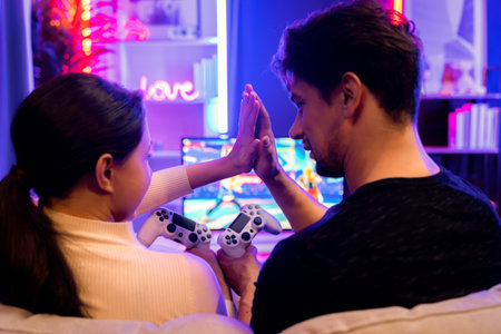 Couple or friend buddy joyful player video game on TV using joystick making hands high five at back side view, winner with blurry screen in studio room in neon light at comfy living home. Postulate.の写真素材
