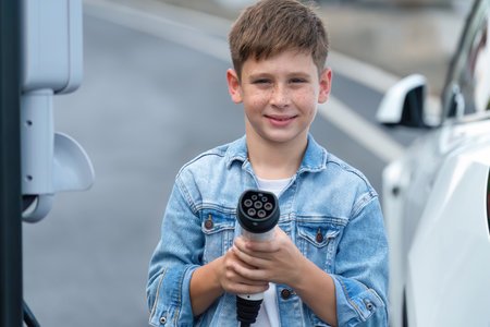 Road trip vacation with eco-friendly EV car and little boy pointing EV charger at camera. Electric...