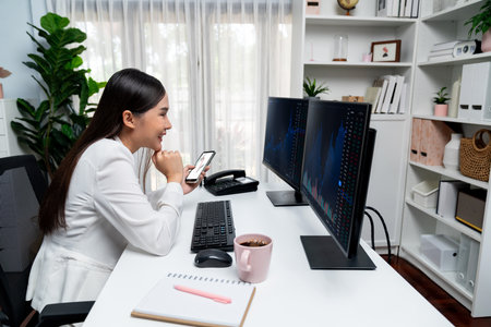 Smiling young Asian businesswoman with happy face looking pc comparing with smartphone in profitable of exchange stock market graph at modern office investing high profit wealth security. Stratagem.の写真素材