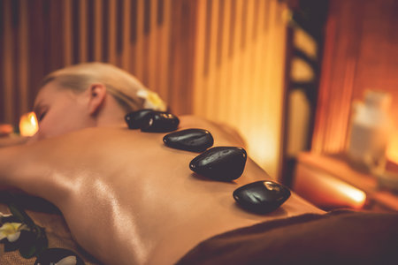 Closeup hot stone massage at spa salon in luxury resort with warm candle light, blissful woman customer enjoying spa basalt stone massage glide over body with soothing warmth. Quiescentの写真素材