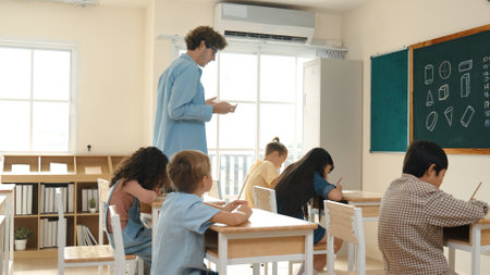 Skilled teacher walking at classroom while looking at student homework. Professional instructor checking smart children notebook while student doing test or working on homework at classroom. Pedagogy.の写真素材