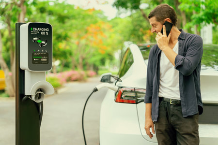 Man talking on smartphone while recharging electric car battery charging from EV charging station during vacation holiday road trip at national park or autumnal forest. Exaltの写真素材