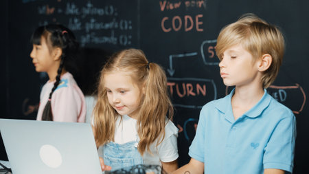 Smart girl using laptop programing engineering code with diverse friends ay table with microchip and main board scatter around in STEM classroom at blackboard written with coding prompt. Erudition.の写真素材