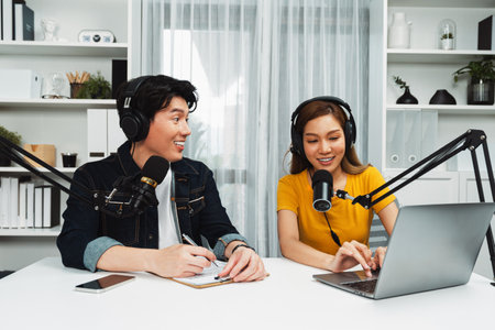 Smiling radio influencer hosts wearing headphones with channel social media online live streaming with talking topic on script note and laptop to listeners at morning time at studio record. Infobahn.の写真素材