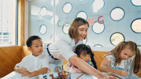 Teacher clicking at laptop while diverse student looking at screen. Children excited in presentation while drawing or writing art books at table with toys and color pencil placed on table. Erudition.の写真素材