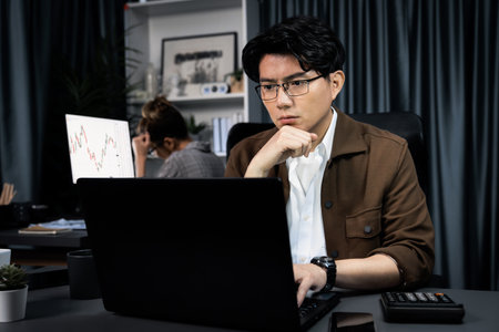 Serious face of thinking businessman focusing laptop to report data analysis social media online project with coworker analyzing stock at back side at over late night time at modern office. Infobahn.の写真素材
