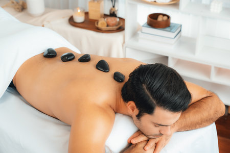 Hot stone massage at spa salon in luxury resort with day light serenity ambient, blissful man customer enjoying spa basalt stone massage glide over body with soothing warmth. Quiescentの写真素材
