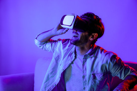 Happy man sitting at sofa while wearing VR goggle to watch funny movie. Caucasian person relaxed...