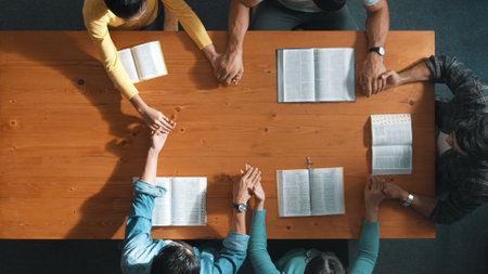Top view of prayer hold each other support while prays to god at table with bible and cross placed...