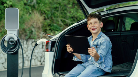 Little boy sitting on car trunk, using tablet while recharging eco-friendly car from EV charging station. EV car road trip travel as alternative vehicle using sustainable energy concept. Perpetualの写真素材