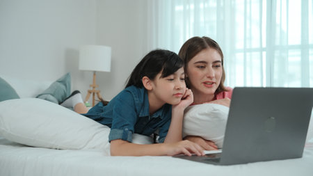 Attractive daughter and caucasian mother watching movie at laptop screen while lie on bad. Happy...