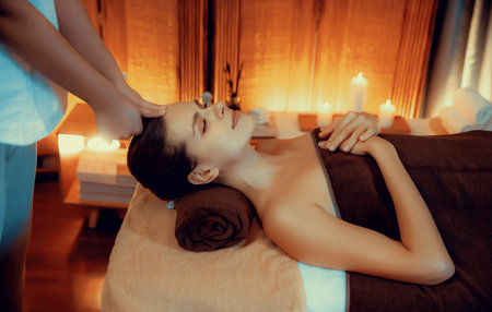 Caucasian woman enjoying relaxing anti-stress head massage and pampering facial beauty skin recreation leisure in warm candle lighting ambient salon spa in luxury resort or hotel. Quiescentの写真素材