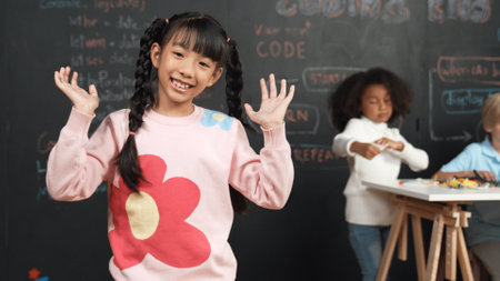 Girl smile and wave to camera while friend working or learning engineering code or prompt in STEM technology classroom. Student looking at camera and greeting while children using laptop. Erudition.の写真素材