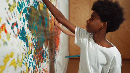 Back view of happy african boy painted the stained wall with colorful hand while wearing casual white shirt in art lesson.Smart student use hand print to make creative artwork. Education. Edification.の写真素材