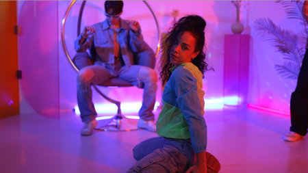 Attractive happy dancer looking at camera while smart woman moving at rhyme with neon light....