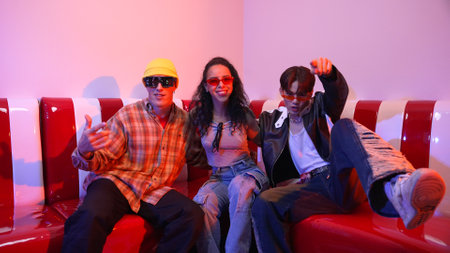 Happy girl looking at camera and moving to pop music while sitting at sofa restaurant. Hispanic woman staring at camera while dancing to freestyle beat with diverse friend in neon light. Regalement.の写真素材