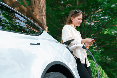 Young woman use smartphone to pay for electricity at public EV car charging station green city park. Modern environmental and sustainable urban lifestyle with EV vehicle. Expedientの写真素材