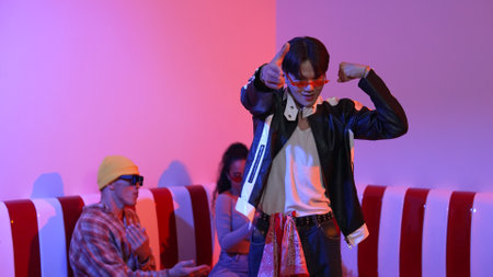 Asian man standing and dancing with pop music while multicultural friend sitting behind at home with led light. Attractive street dancer moving to hip hop music while looking at camera. Regalement.の写真素材