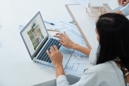 Beautiful caucasian architect using laptop while smart coworker measures house model with blueprint, equipment placed on table. Creative business design and teamwork concept. Top view. Immaculate.の写真素材