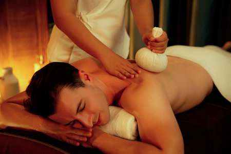 Hot herbal ball spa massage body treatment, masseur gently compresses herb bag on man body. Tranquil and serenity of aromatherapy recreation in warm lighting of candles at spa salon. Quiescentの写真素材