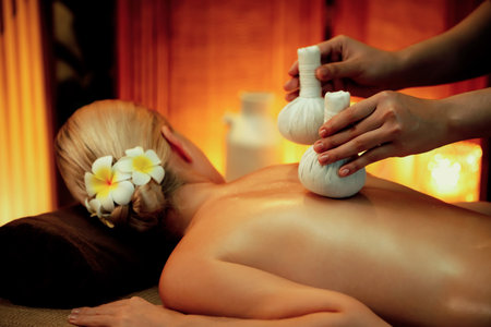Hot herbal ball spa massage body treatment, masseur gently compresses herb bag on woman body. Tranquil and serenity of aromatherapy recreation in warm lighting of candles at spa salon. Quiescentの写真素材