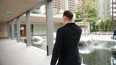 Back view of happy manager talking to phone while walking at modern corridor. Smart boss using telephone by calling marketing team to plan about financial strategy while walking at green city. Urbane.の写真素材