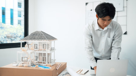 Closeup image of professional architect engineer using laptop analysis data on meeting table with house model and map scatter around at room with blueprint hanged behind. Focus on hand. Immaculate.の写真素材