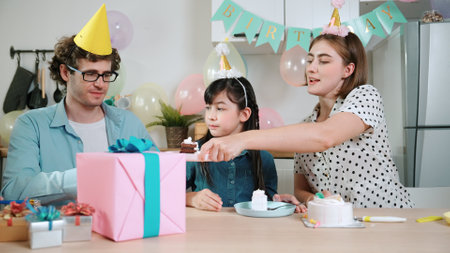 Caucasian mom cutting birthday cake and separated dessert while give to father. Happy family wear party hat to celebrated daughter birthday while sitting at table. Special occasion concept. Pedagogy.の写真素材