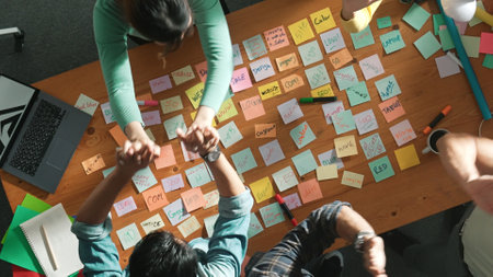 Top view of business team giving high five to celebrate project while standing at table with sticky notes and laptop. Aerial view of designer clapping hands to congratulate successful idea. Symposium.の写真素材